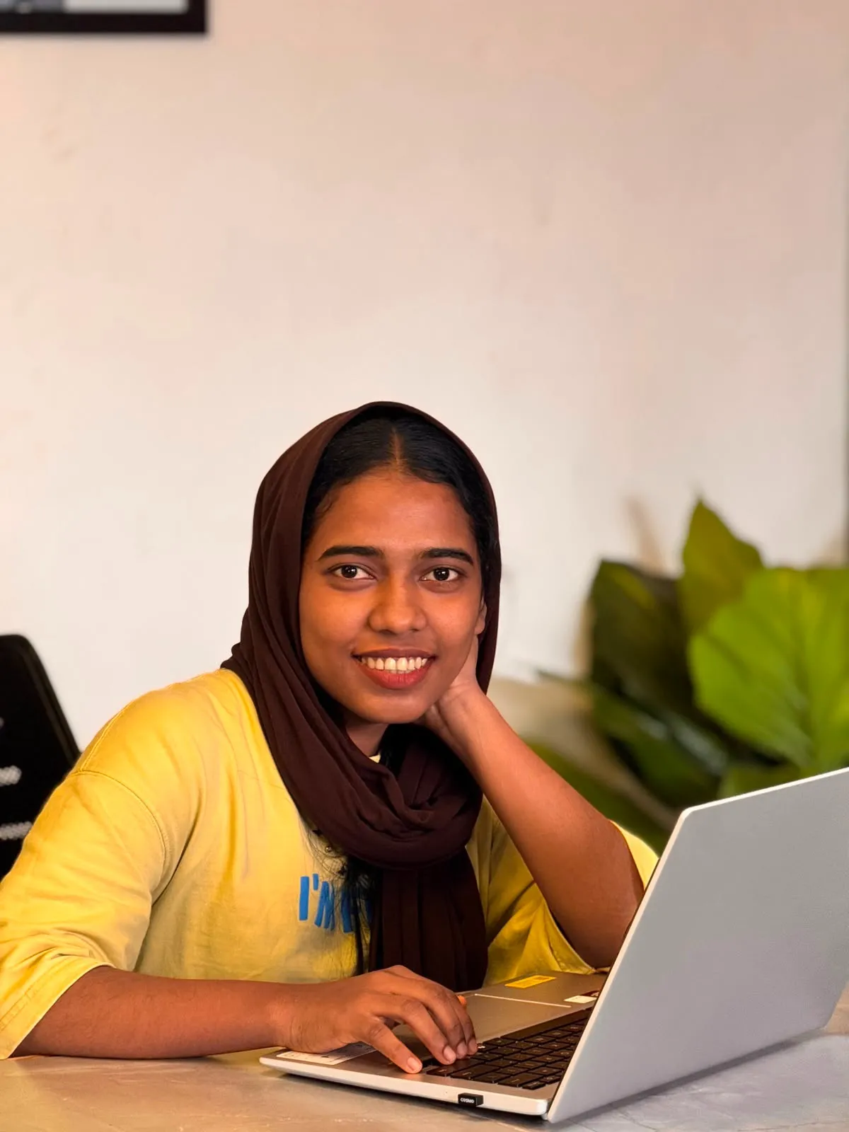 A woman wearing a brown hijab and yellow top sits at a desk using a laptop, smiling at the camera in an indoor setting with a plant in the background